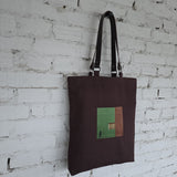 Brown tote bag with a green and brown square design against a white brick wall.
