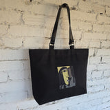 A black canvas tote bag with black shoulder straps, featuring a stylized cubist face graphic in yellow, gray, and black on the front, hanging against a white painted brick wall.