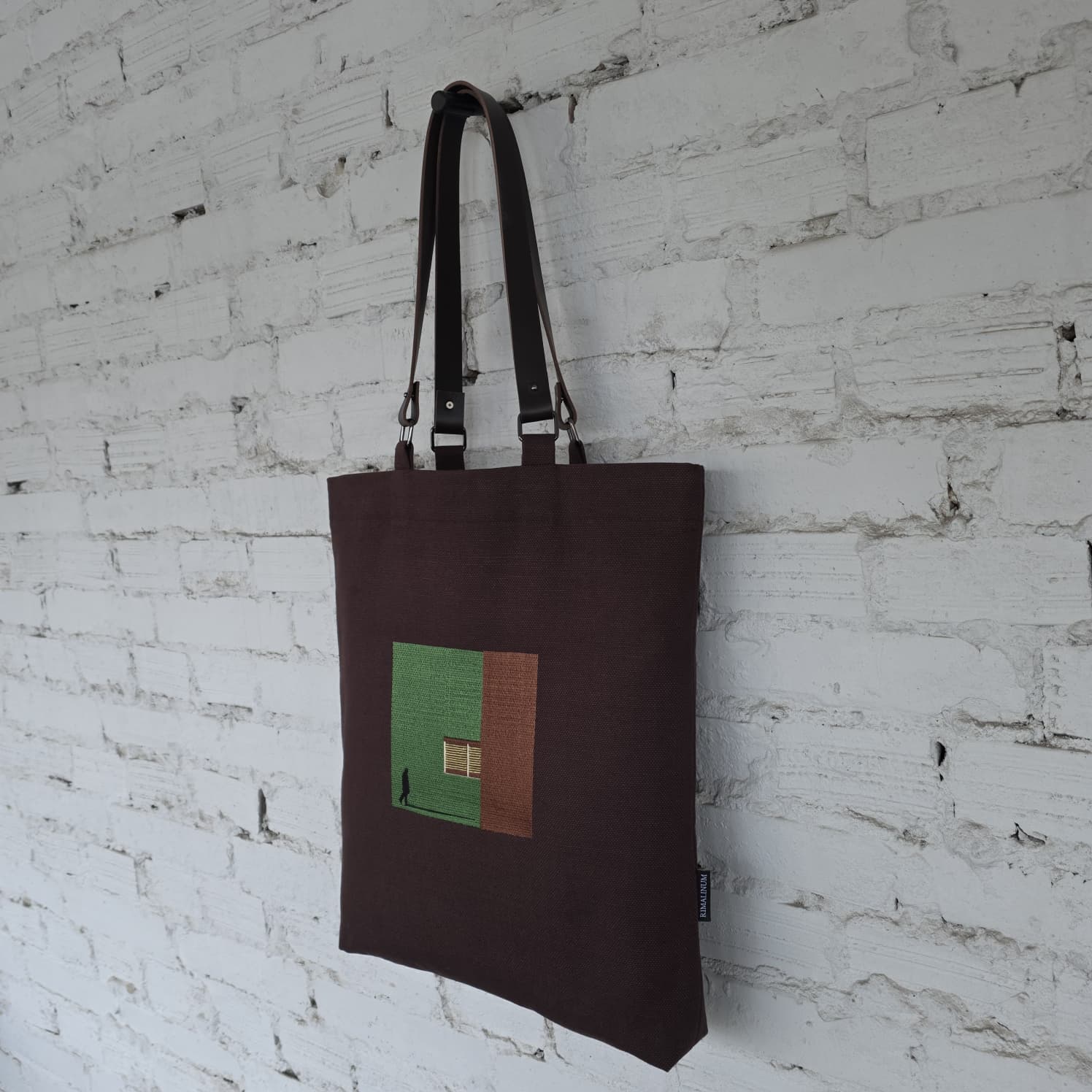 Brown tote bag with a green and brown square design against a white brick wall.