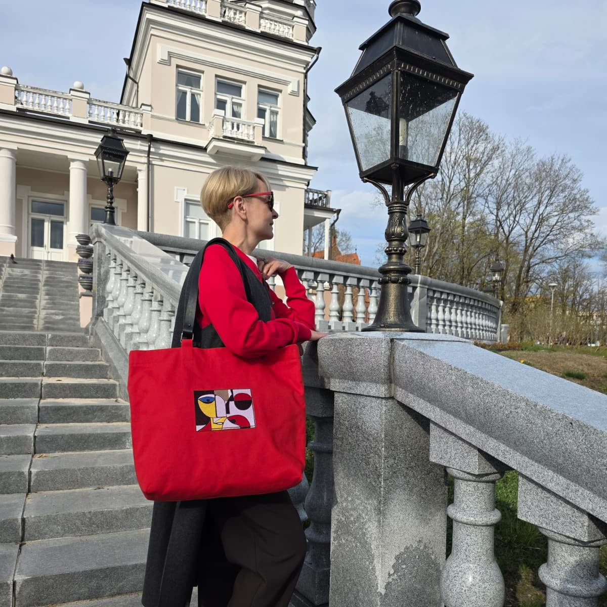 A woman with short blonde hair and sunglasses stands on stone stairs carrying a large red tote bag with a colorful graphic print. She is wearing a red top, black coat, and black pants, with a grand building and a black street lamp in the background.