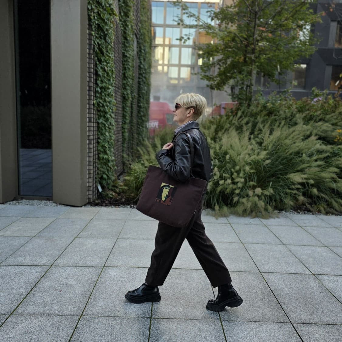 A person with short blonde hair and sunglasses walks across a paved outdoor area, wearing a black leather jacket, dark pants, and carrying a large dark brown tote bag.
