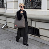 A woman walking on a city sidewalk carrying a large khaki hobo bag with a small red and grey embroiderd design. She is wearing a black top, dark wide-leg plaid trousers, and black shoes, standing in front of a light gray stone building.