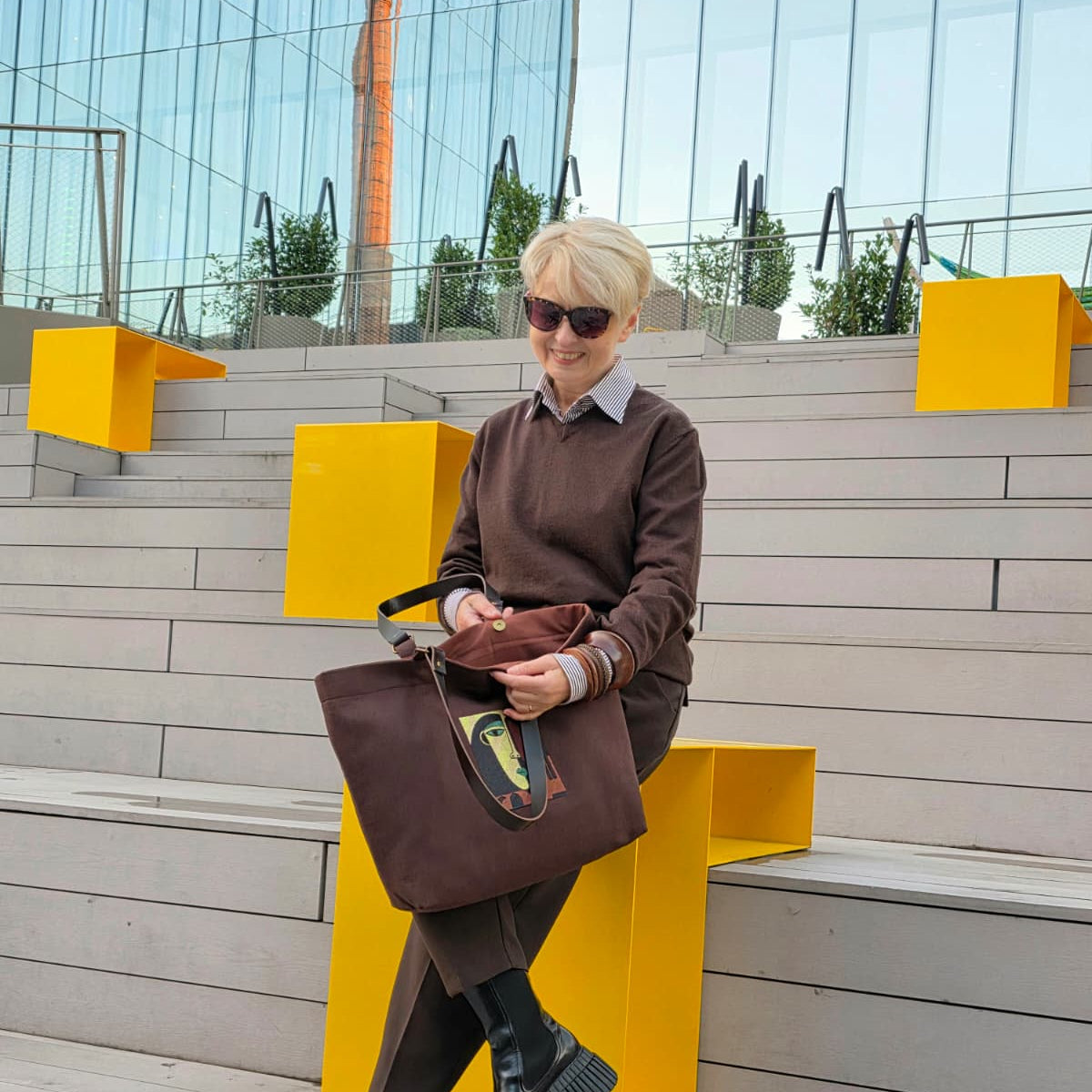 A woman with short blonde hair and sunglasses sits on a modern outdoor concrete bench with bright yellow geometric accents. She is wearing a brown sweater, brown trousers, and black chunky boots, holding a large brown leather tote bag .