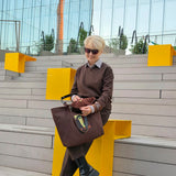 A woman with short blonde hair and sunglasses sits on a modern outdoor concrete bench with bright yellow geometric accents. She is wearing a brown sweater, brown trousers, and black chunky boots, holding a large brown leather tote bag .