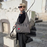 A woman with short blonde hair and sunglasses stands on outdoor stone steps, wearing a large khaki hobo bag with a small red and grey embroidered design over her shoulder.