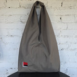 A khaki waterproof hobo bag  with a small red, black, and grey ebroidered design on the lower left, hanging on a black stand against a white brick wall. 