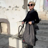 A woman with short blonde hair and sunglasses stands on outdoor stone steps holding a large khaki hobo  bag . She is wearing a black top and dark pants, with a small red-and-grey embroidered design near the bottom of the bag.