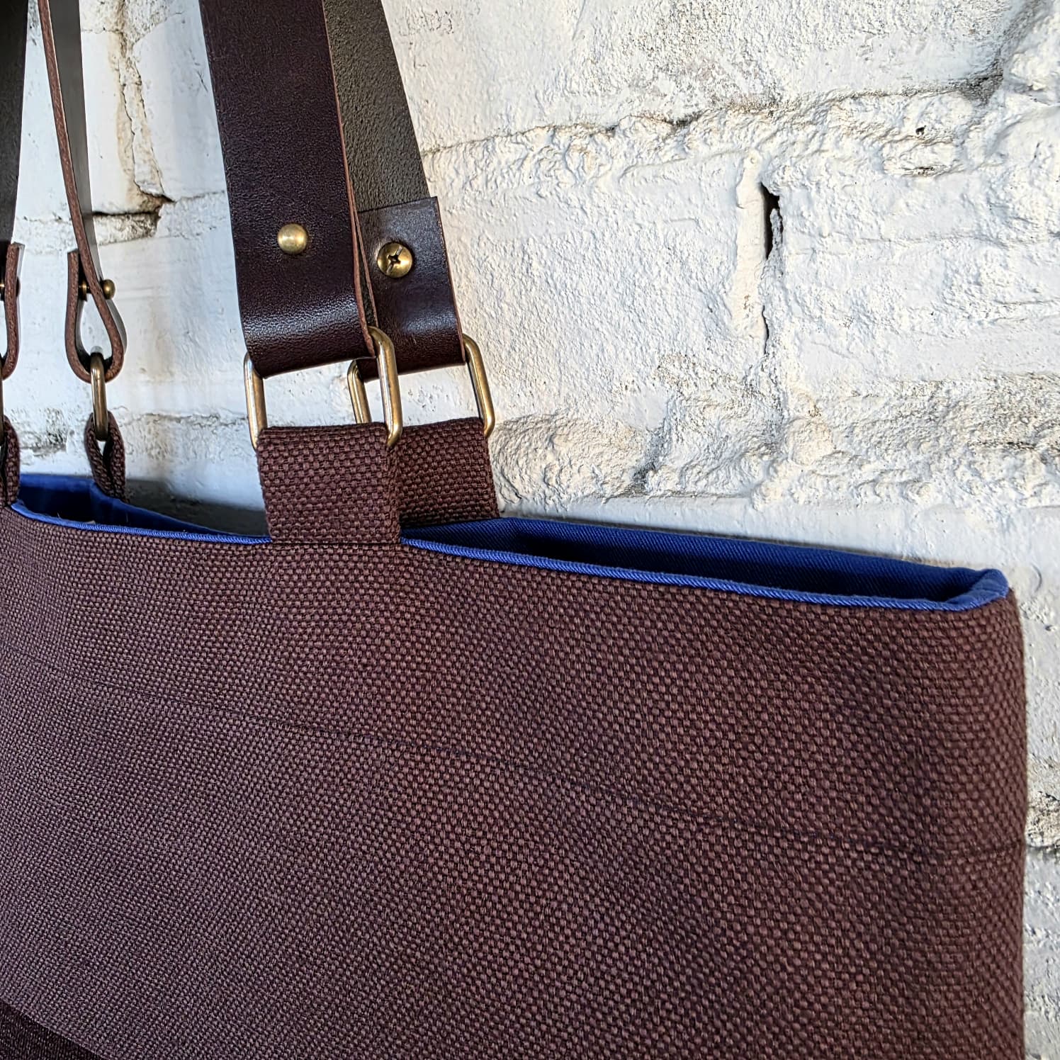 A close-up of a brown woven tote bag with dark brown leather shoulder straps and brass hardware, shown against a white painted brick wall.
