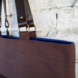 A close-up of a brown woven tote bag with dark brown leather shoulder straps and brass hardware, shown against a white painted brick wall.